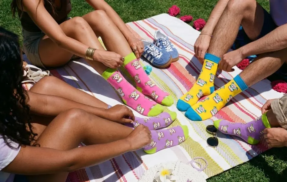 people-wear-socks A group of friends sitting together on a picnic blanket on the lawn, showcasing yellow, pink, and purple Gary the Fire Goat crew socks.